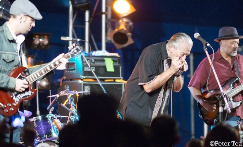 Three people on stage, one playing a harmonica, two playing guitar