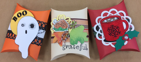 Three pillow box gift holders. An orange and black with a ghost and the word BOO; one tan and brown with a picture of a cooked turkey and the word GRATEFUL; one is red with a green holly branch and cup of hot chocolate with marshmallows