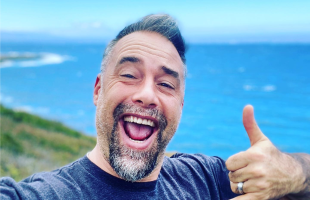 Man in blue shirt with ocean behind smiling and holding up a shaka