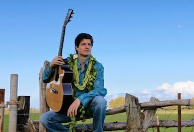 musician Jeff Peterson, holding a guitar while sitting on a fence outdoors.