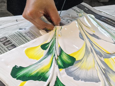 Hand pulling string through paint to create flower shapes on canvas in green, white, and yellow.