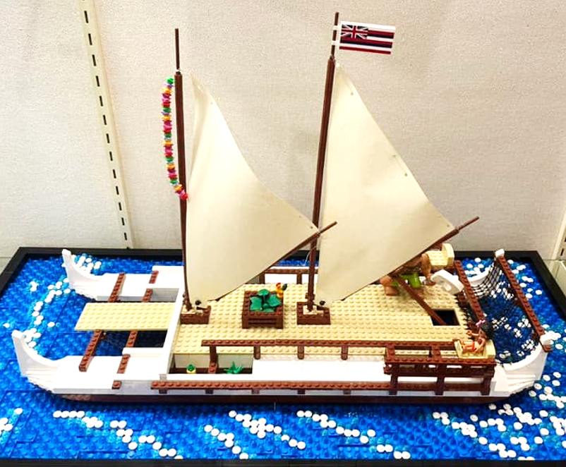 A double hulled canoe with two sails, one flying the Hawaiian flag, on a blue-and-white base that mimics water and seafoam, all created out of LEGO pieces.