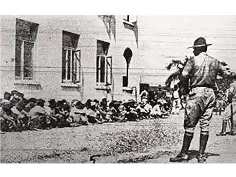 Black & White Image of National guard soldiers watching over 130 strikers awaiting trial for riot charges outside the Lihue court.