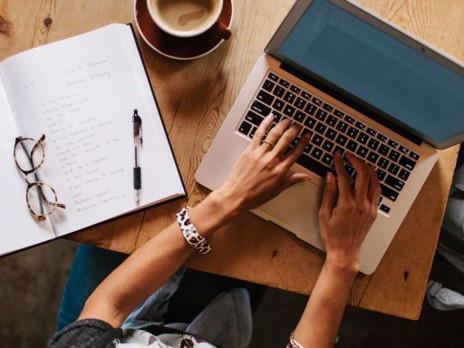 Hands typing on a typewriter with tablet, glasses, pen and coffee mug nearby.