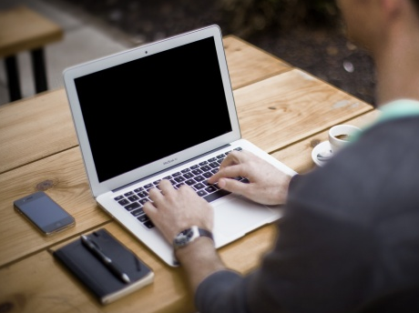 A laptop sitting on a wooden table with a smart phone and a datebook and pen to the left of it. A man is typing on the laptop.