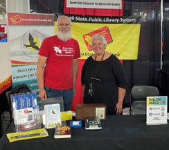 A man in a red shirt and a woman in a black shirt standing behind a display table