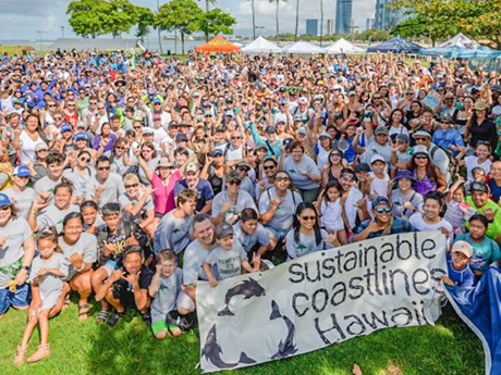 Sea of people gathered at a park. Those in the front of the photo hold a banner that reads "Sustainable Coastlines Hawaiʻi".