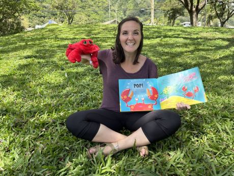 A woman holding a book and crab plushy is sitting on grass at a park