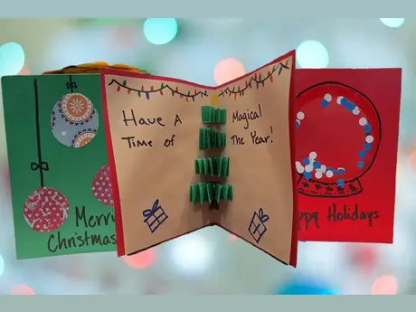 A picture of three homemade holiday cards: a green card with the words Merry Christmas, a tan card with a 3D Christmas tree with the words have a magical time of the year, and a red card with a snow globe on it that says happy holidays