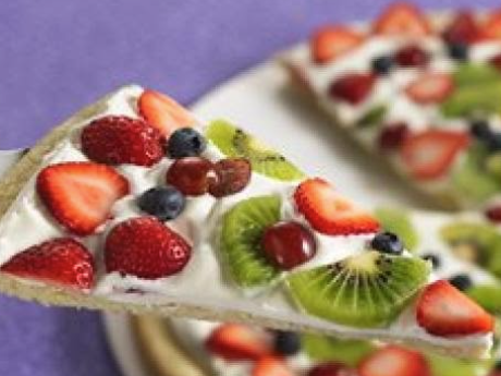 A wedge of cookie topped with cream cheese spread and on top of that sliced strawberries, grapes, and kiwi fruit