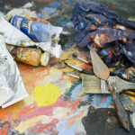 Paints, brushes, and painting knives are strewn across a dirty pallet.