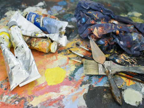 Paints, brushes, and painting knives are strewn across a dirty pallet.