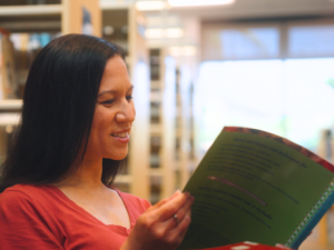 A woman is seated in a library, focused on a book, with numerous bookshelves in the background.