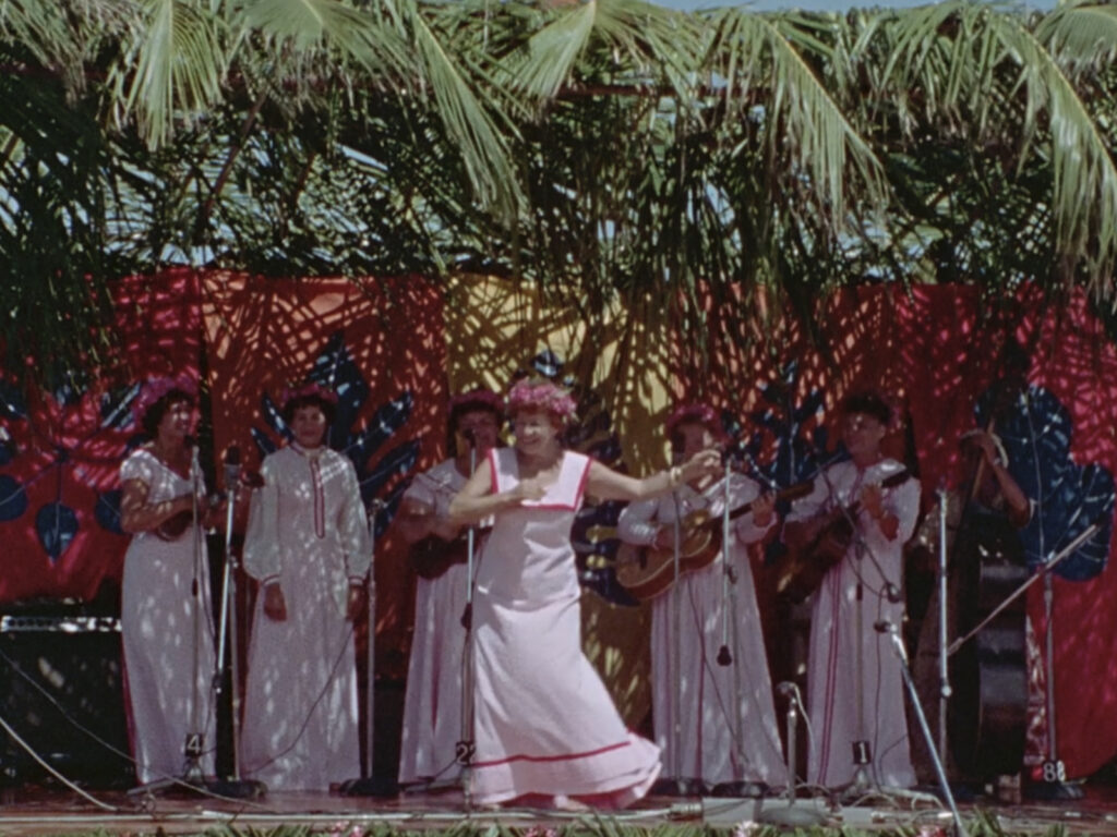 A woman in a white muʻumuʻu dances hula in front of a band of five women playing instruments and singing.