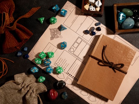 A blank character sheet under a book and multiple sizes of dice and stones with a couple small pouches and trays to the side.