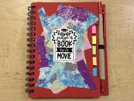 red spiral bound notebook with strips of duct tape and a book-themed sticker on it