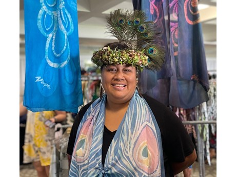 A smiling woman with peacock feathers in her hair and lei po'o on her head, wearing a scarf.