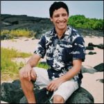 Man sitting on beach rocks, aloha shirt, white shorts, black hair