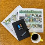 A wooden table holds two folded newspapers, a black notebook with a pen featuring a colorful logo, and a yellow mug filled with coffee.