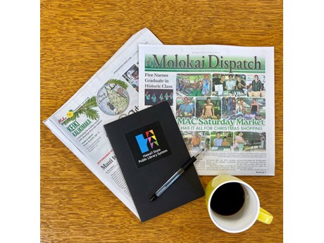 A wooden table holds two folded newspapers, a black notebook with a pen featuring a colorful logo, and a yellow mug filled with coffee.