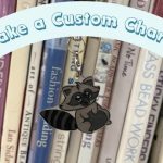 Photo of a racoon design phone charm hanging in front of books. A banner reads "Make a Custom Charm"