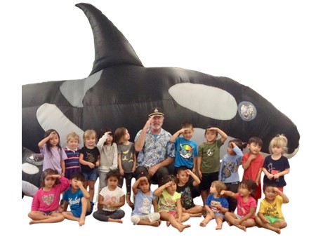 A group of smiling children sits in front of a large inflatable orca. An adult in a captain's hat is seated in the center, surrounded by the kids.