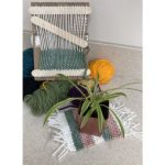 A pile of yarn with a cardboard loom on top. The loom has a half finished project on it. In the foreground there's a miniature woven rug being used as a coaster for a potted plant.