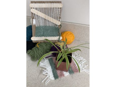 A pile of yarn with a cardboard loom on top. The loom has a half finished project on it. In the foreground there's a miniature woven rug being used as a coaster for a potted plant.
