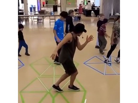A group of children and a leader engage in a lively dance activity on a gym floor, marked with colorful geometric tape shapes
