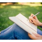 A woman sits on the grass at a park. She's writing in a notebook that's on her lap with a pen.