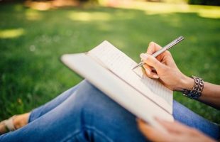 A woman sits on the grass at a park. She's writing in a notebook that's on her lap with a pen.