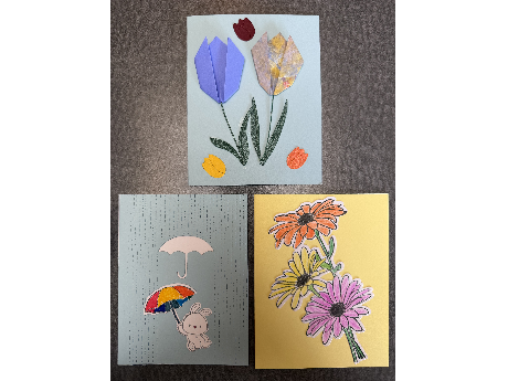 Three handmade cards, one with folded paper tulips, one with stamped and colored daisies, and one with a rabbit in the rain with two umbrellas