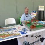 A person sits at an informational booth with educational materials and a 3D model, holding up a sign that reads "ACT 125."