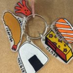 Picture of keychain consisting of shrinky dinks of shrimp tempura, onigiri, bento box, salmon nigiri, and octopus-shaped sausage with
