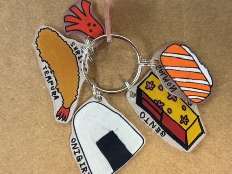 Picture of keychain consisting of shrinky dinks of shrimp tempura, onigiri, bento box, salmon nigiri, and octopus-shaped sausage with
