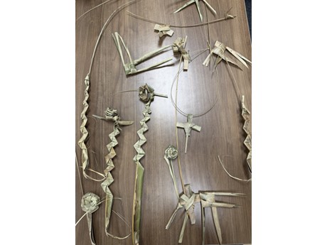 Various intricate palm leaf creations are arranged on a wooden table. The crafts include woven fish and roses.