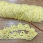 On the brown table, there are an orange crochet hook, light yellow yarn skein and a crocheted crown made out of the same light yellow yarn