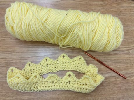 On the brown table, there are an orange crochet hook, light yellow yarn skein and a crocheted crown made out of the same light yellow yarn