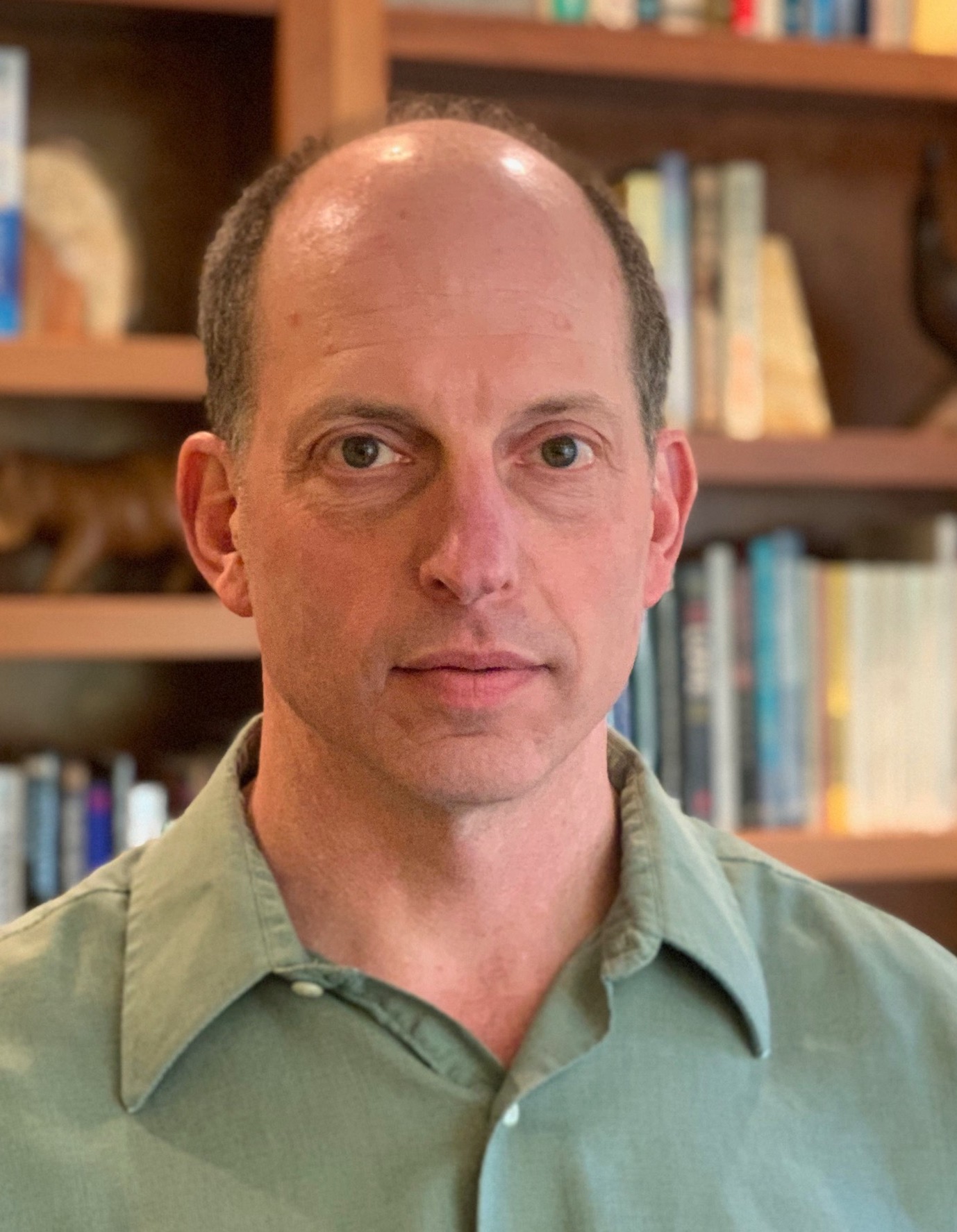 Pictured: headshot of a man in a green shirt in front of a bookcase.