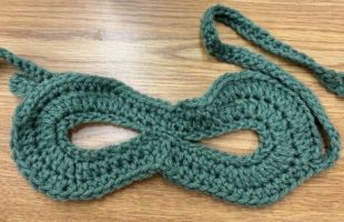 Green crocheted masquerade mask laid flat on brown table.