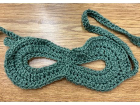 Green crocheted masquerade mask laid flat on brown table.