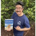 Poet Laureate Lee A. Tonouchi holding the book Chiburu and giving a shaka
