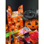 Goodie bag decorated with cats, bats, pumpkins, and “BOO!” lettering, surrounded by assorted candy and next to a jack-o'-lantern container filled with black roses.
