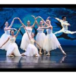 6 female and 1 male ballet dancers wearing white costumes on stage in front of blue image background