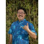 Photo of a man with black hair, a mustache, and glasses wearing a blue aloha shirt and making a shaka sign