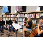 Four musicians play a cello and violin in a library, seated among music stands and bookshelves; the focused scene feels calm and collaborative.